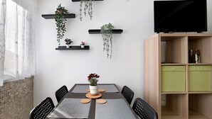 Een eetgedeelte met een tafel voor vier personen, omringd door stoelen, een aan de muur bevestigde televisie en een boekenkast met groene kussens.