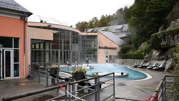 Zona de piscina con un edificio de vidrio, sillas de descanso y un paisaje agreste de colinas.