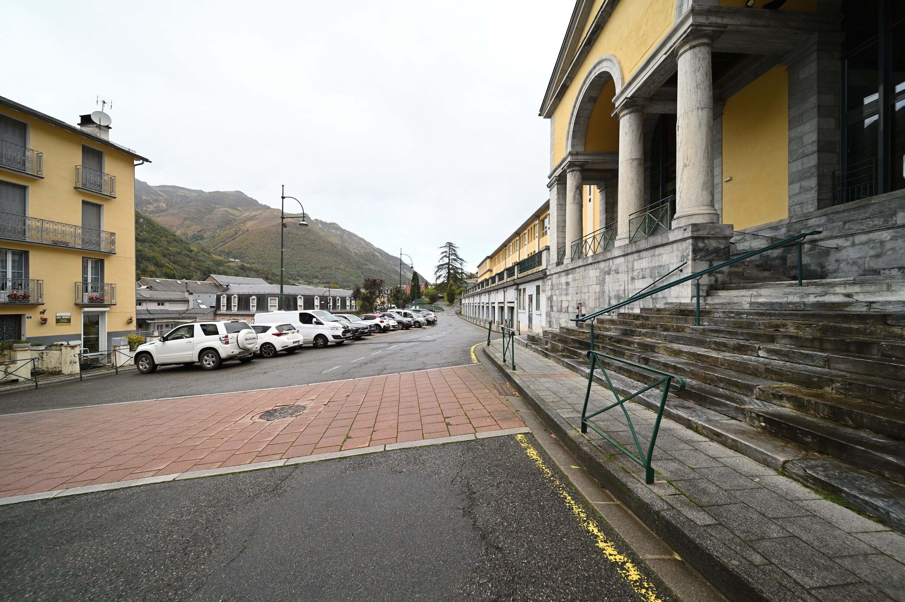 A street with parked cars, a building with columns, and a mountain in the background.