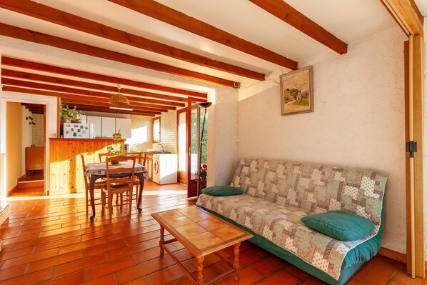 A cozy living room with a wooden floor, a sofa with a quilt, a wooden coffee table, and a dining area with chairs and a table.
