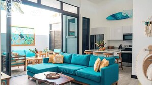 A modern living room with a teal sectional sofa, a wooden coffee table, and a kitchen area with white cabinets and a large window.