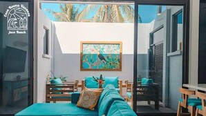 A modern living room with a teal sofa, wooden chairs, and a painting of a tropical scene.