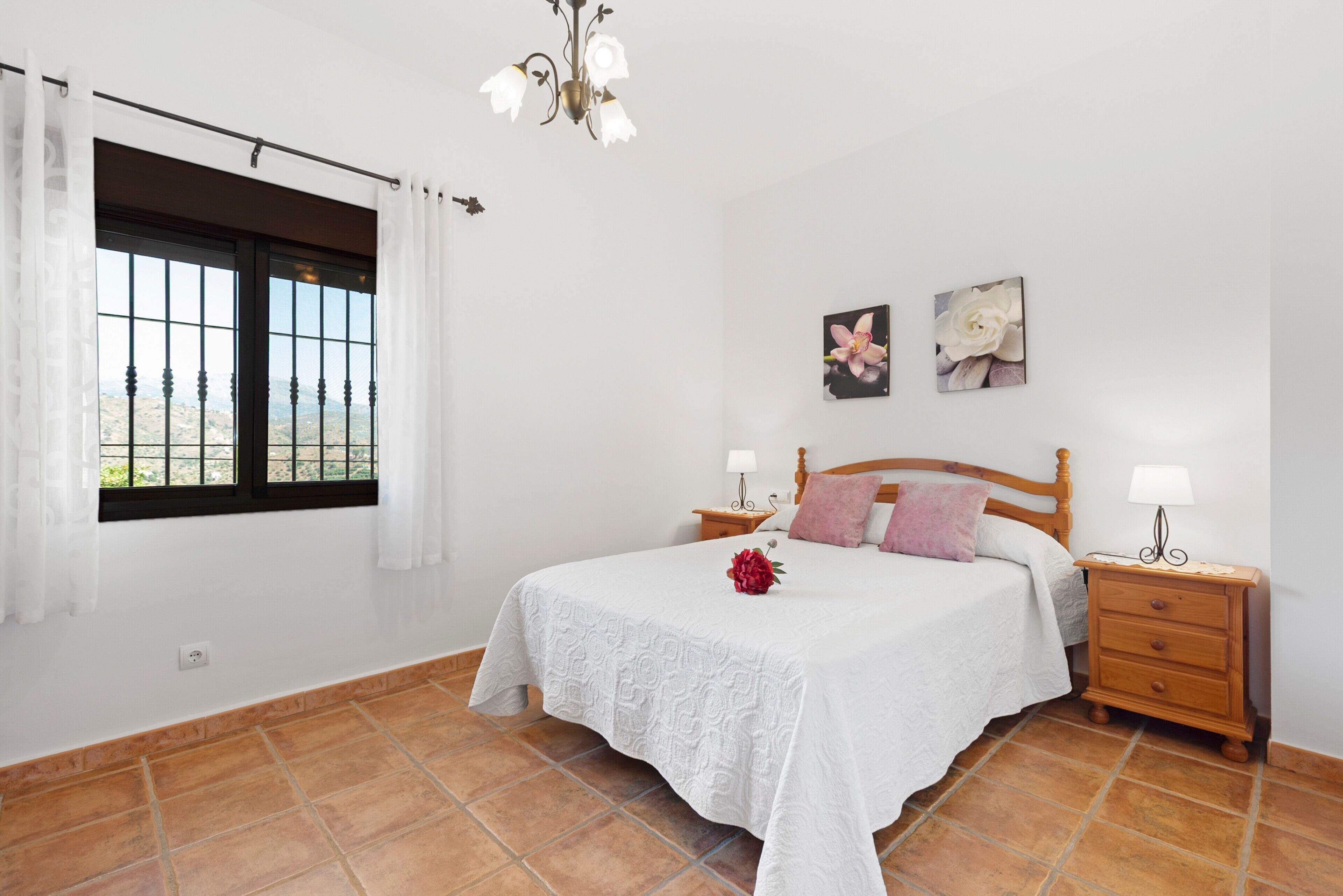 Une chambre à coucher comprenant un lit, deux tables de chevet, une fenêtre avec des rideaux, un lustre et deux tableaux représentant des fleurs, accrochés au mur.