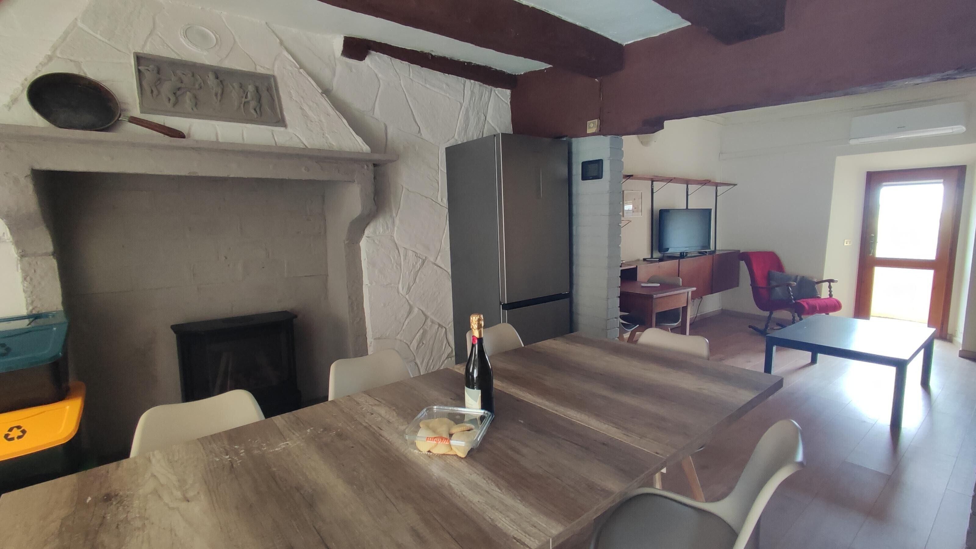 Ein Essbereich mit Holztisch, Stühlen, einer Weinflasche, einem Kamin, einem Fernseher und einem roten Sessel.