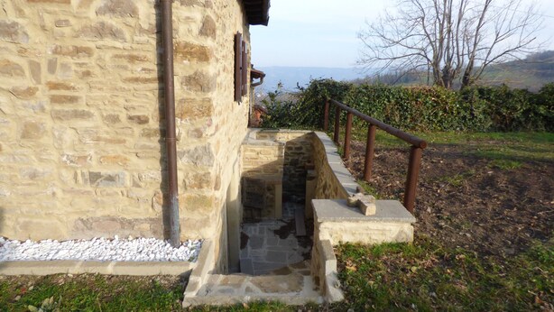 A stone building with a narrow stone staircase leading to a door, surrounded by greenery and a view of the landscape.