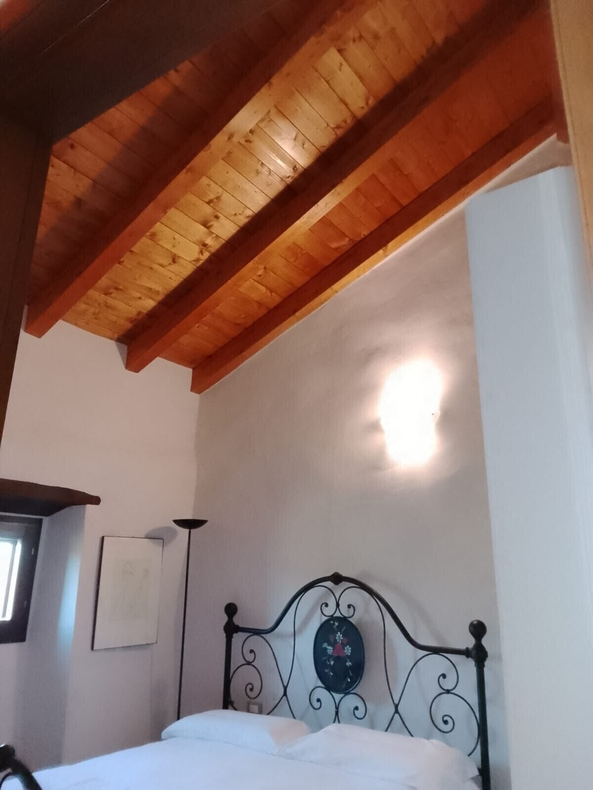Une chambre avec un plafond en bois, un lit doté d’une tête de lit en métal, un luminaire fixé au mur et un tableau encadré.