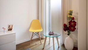 Une pièce avec une table en verre, une chaise jaune, un vase blanc rempli de fleurs et un tableau encadré posé sur une commode blanche.