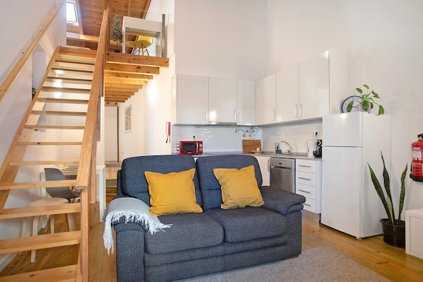 Une cuisine moderne avec des meubles blancs, un escalier en bois, un canapé gris avec des coussins jaunes et une plante en pot.