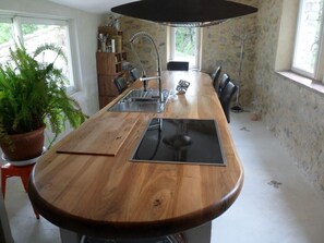 Une longue table en bois, dotée d’un évier intégré, entourée de chaises, dans une pièce aux murs de pierre et à la grande fenêtre.