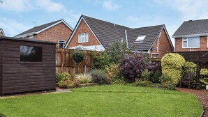 Un jardin résidentiel avec une pelouse, un abri en bois et une maison en briques surmontée d’un toit en tuiles.