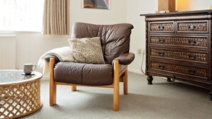 Un fauteuil en cuir marron avec un coussin à motifs, une table d’appoint en bois avec une tasse, une commode en bois avec des tiroirs et une affiche accrochée au mur.