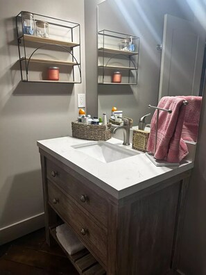 A bathroom with a white sink, wooden vanity, and a mirror above it. There are shelves with toiletries and a pink towel hanging on the wall.