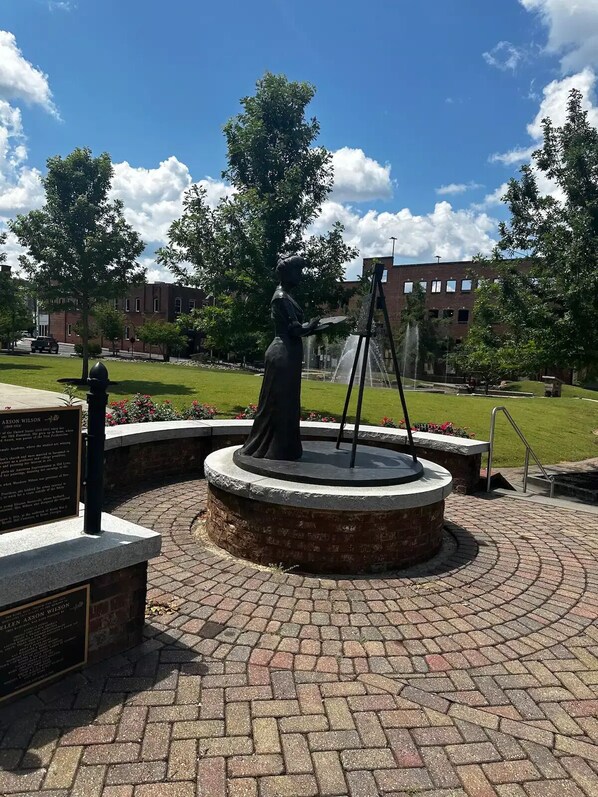 A statue of a woman painting on an easel in a circular brick plaza with a fountain, surrounded by trees and a building in the background.
