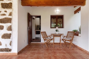 A patio with two wooden chairs, a small round table, a window with a plant, and a stone wall.