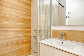 A bathroom with a wooden wall, a glass shower enclosure, a white sink, and a mirror. - Apartment 'La Dune De Sable Bonne Source' with Private Terrace and Wi-Fi (Pornichet)