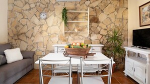 Un salon avec un mur en pierre, un climatiseur blanc, un canapé blanc, une table à manger avec des chaises, un téléviseur et une plante en pot.