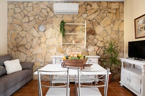 A living room with a stone wall, a white air conditioner, a white sofa, a dining table with chairs, a television, and a potted plant.