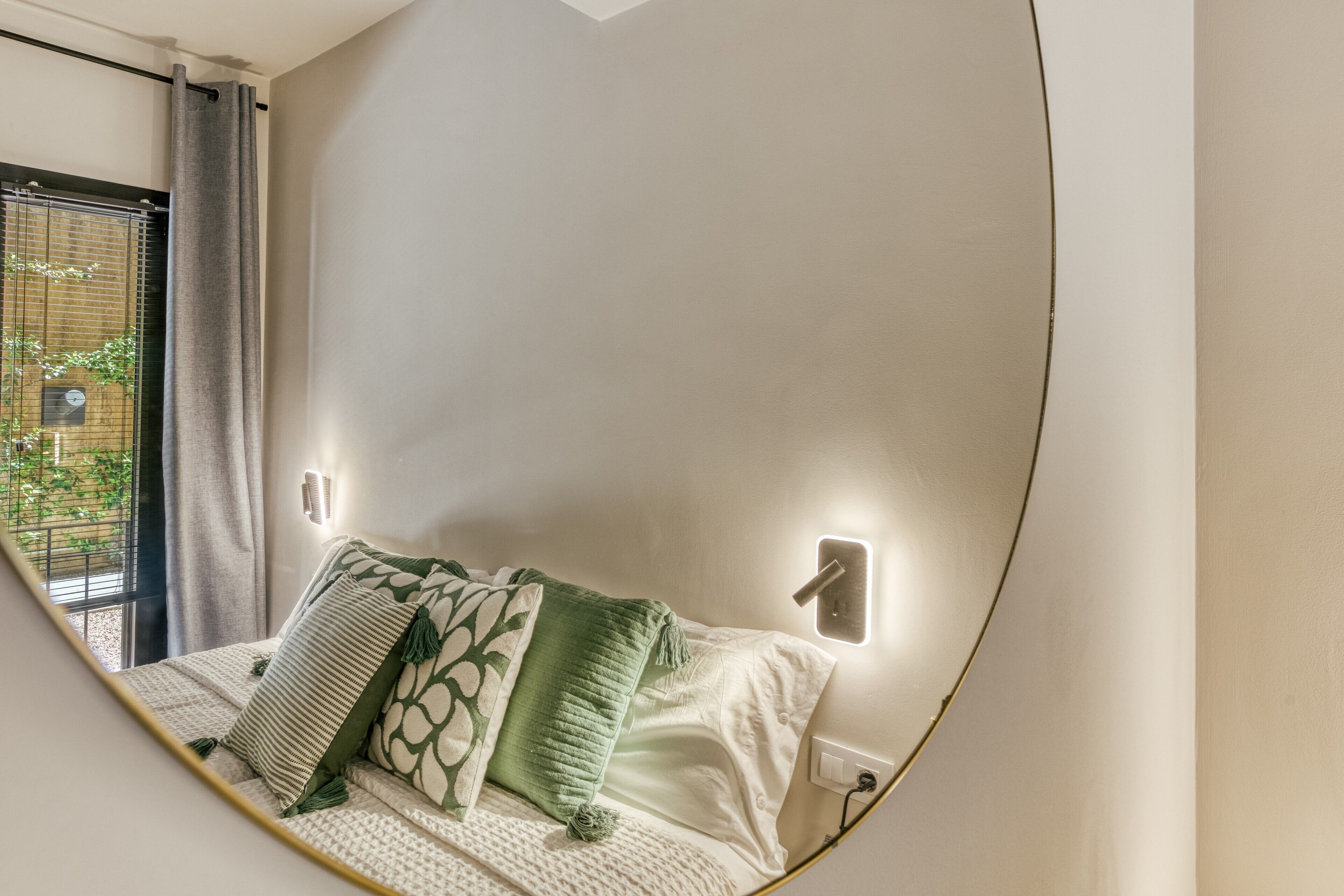 Un dormitorio con cama, almohadas, una lámpara de pared y una ventana con persianas.
