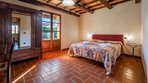 A bedroom with a wooden ceiling, a bed with a patterned bedspread, a bench, and a window with a view of greenery.