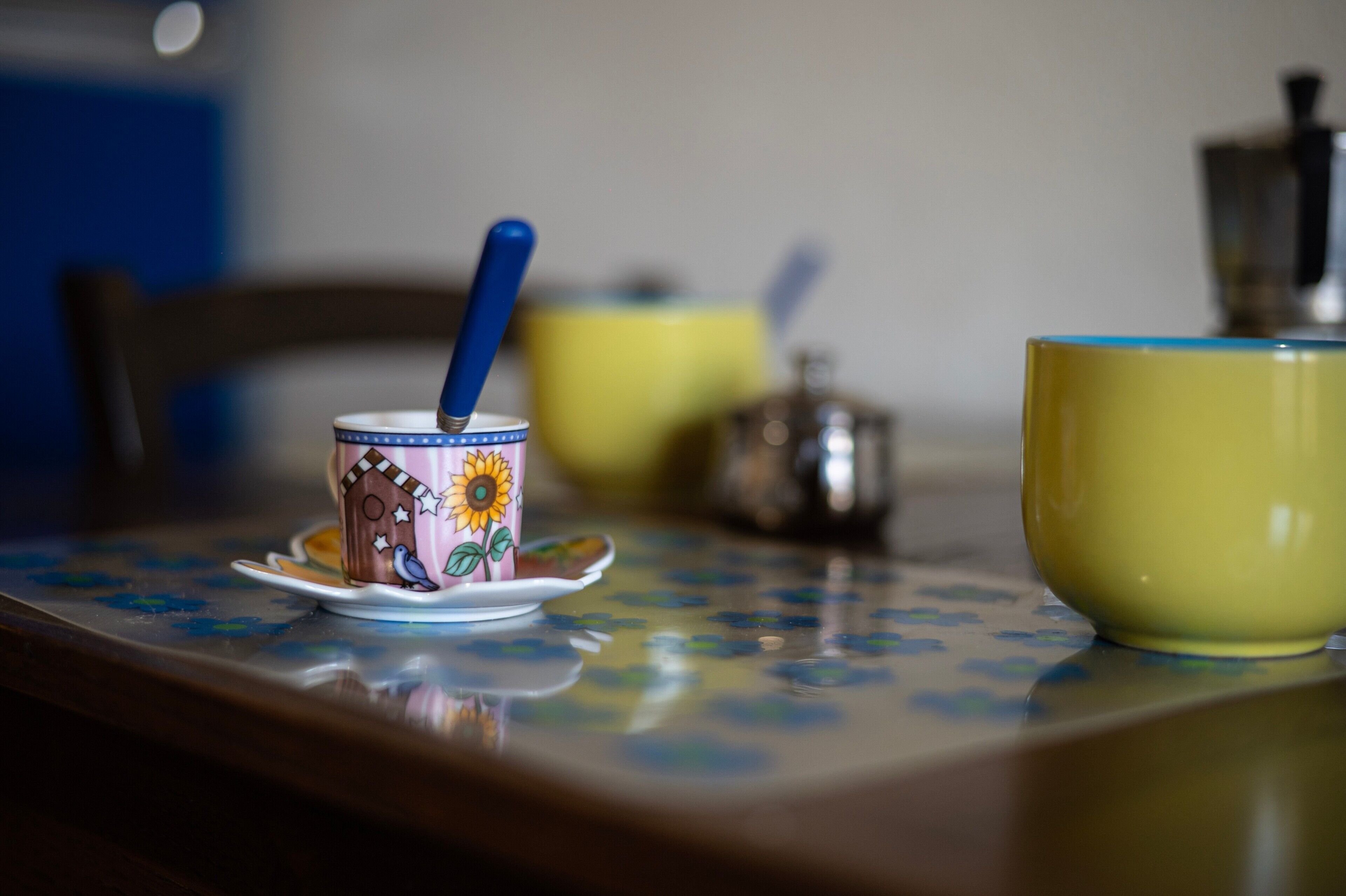 A table with a floral tablecloth, a ceramic mug with a blue handle, a yellow cup, and a small pot.