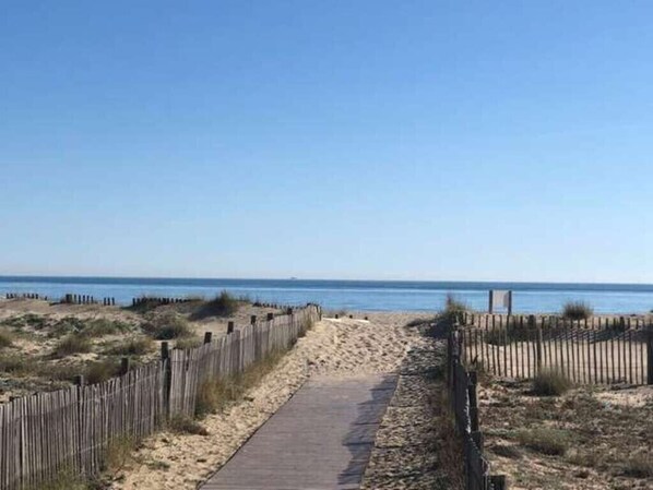 Un sentier de plage sablonneux, bordé d’une clôture en bois d’un côté, et offrant une vue dégagée sur l’océan.