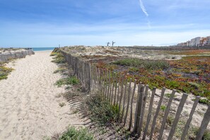 Un sentier de sable bordé d’une clôture en bois, qui mène à une zone de dunes à la végétation clairsemée, d’où l’on a une vue dégagée sur l’océan.