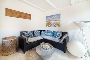 Een hoek zithoek met een donkere rieten bank, gekleurde kussens, een gevlochten mand, een metalen kom op een ronde tafel, een schilderij van een strand en een witte vloerlamp.