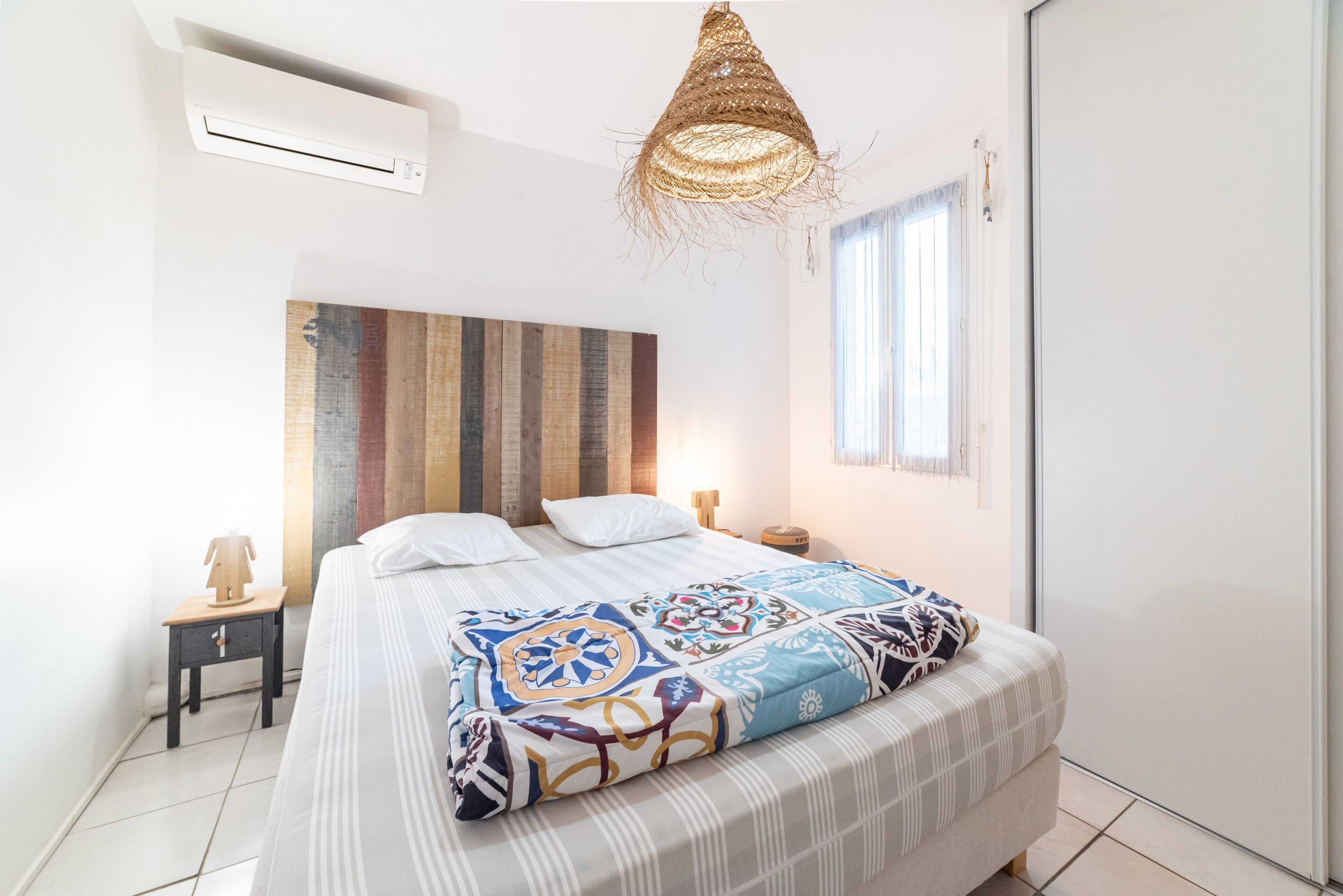 Un dormitorio con cama, aire acondicionado de pared, lámpara colgante de fibras naturales, mesita de noche con lámpara y ventana con persianas.