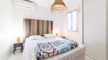 Un dormitorio con cama, aire acondicionado de pared, lámpara colgante de fibras naturales, mesita de noche con lámpara y ventana con persianas.