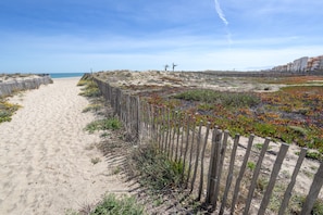 Un sendero arenoso bordeado por una valla de madera, que conduce a una zona de dunas con vegetación escasa y una vista distante del mar.