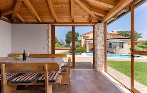 Un comedor de madera con una mesa y bancos, ventanas amplias y vistas a una piscina y un edificio de piedra.