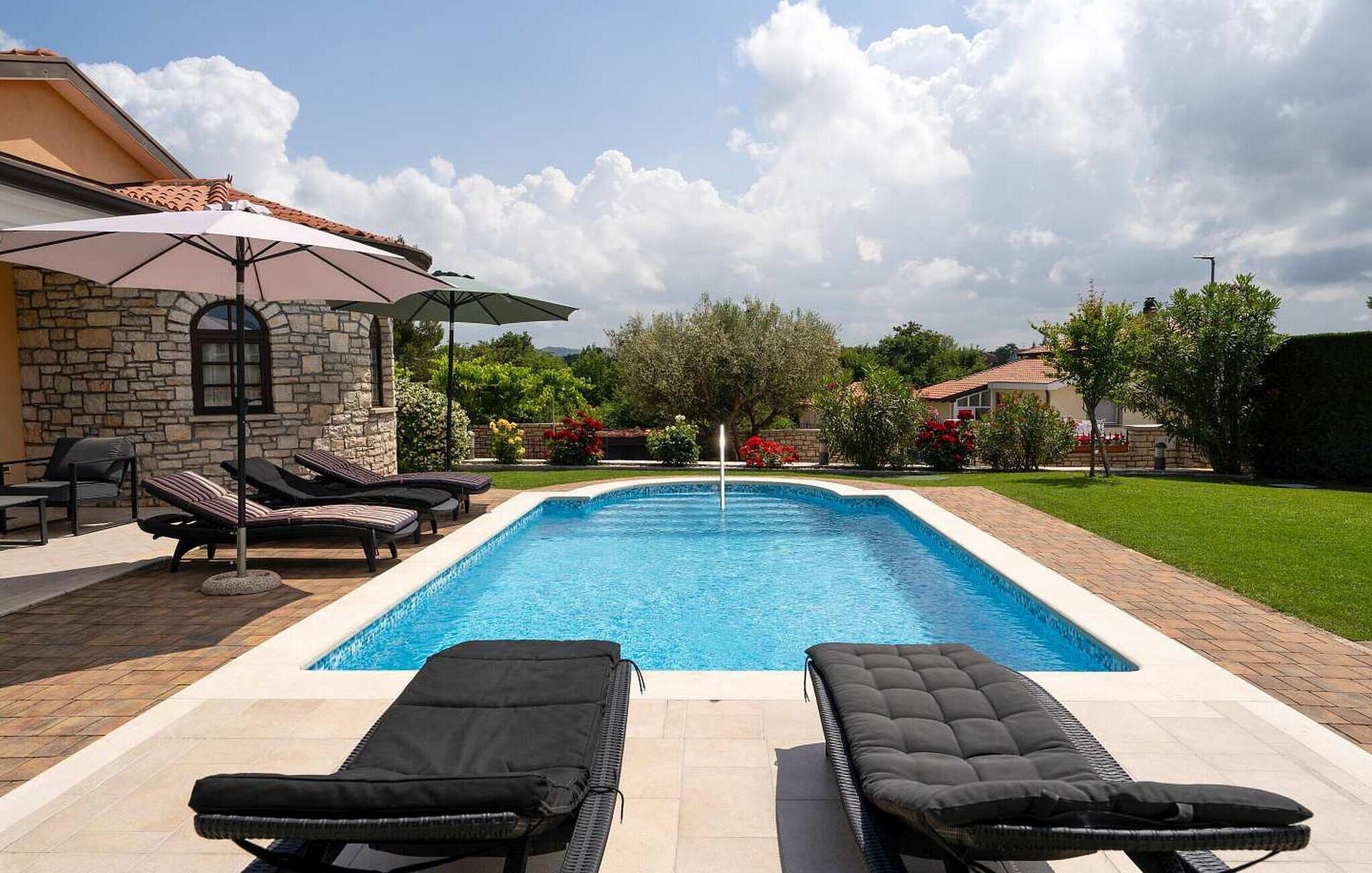 Ein Pool mit Liegestühlen und Sonnenschirm, umgeben von einem Steinhaus und viel Grün.