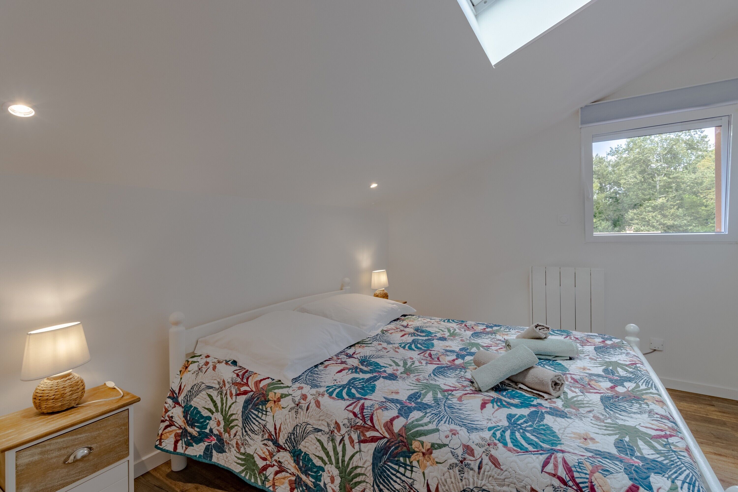 A bedroom with a bed, bedside lamps, a nightstand, a window, and a skylight.