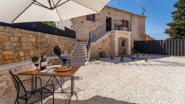 Ein Steinhaus mit einer Treppe, die zu einem Balkon führt. Auf dem Balkon steht ein Tisch mit Pizza und Getränken, und ein Sonnenschirm spendet Schatten.