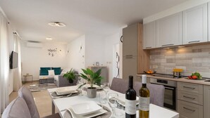 Eine moderne Küche mit hellen Holzschränken, eine Essecke mit einem weißen Tisch für zwei Personen und ein Wohnzimmer mit einem grauen Sofa und türkisfarbenen Kissen.