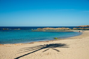 Una playa con aguas azules cristalinas, afloramientos rocosos y algunas personas caminando por la orilla.