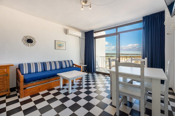 A room with a checkered floor, a sofa with striped cushions, a white table, and a balcony with a view.