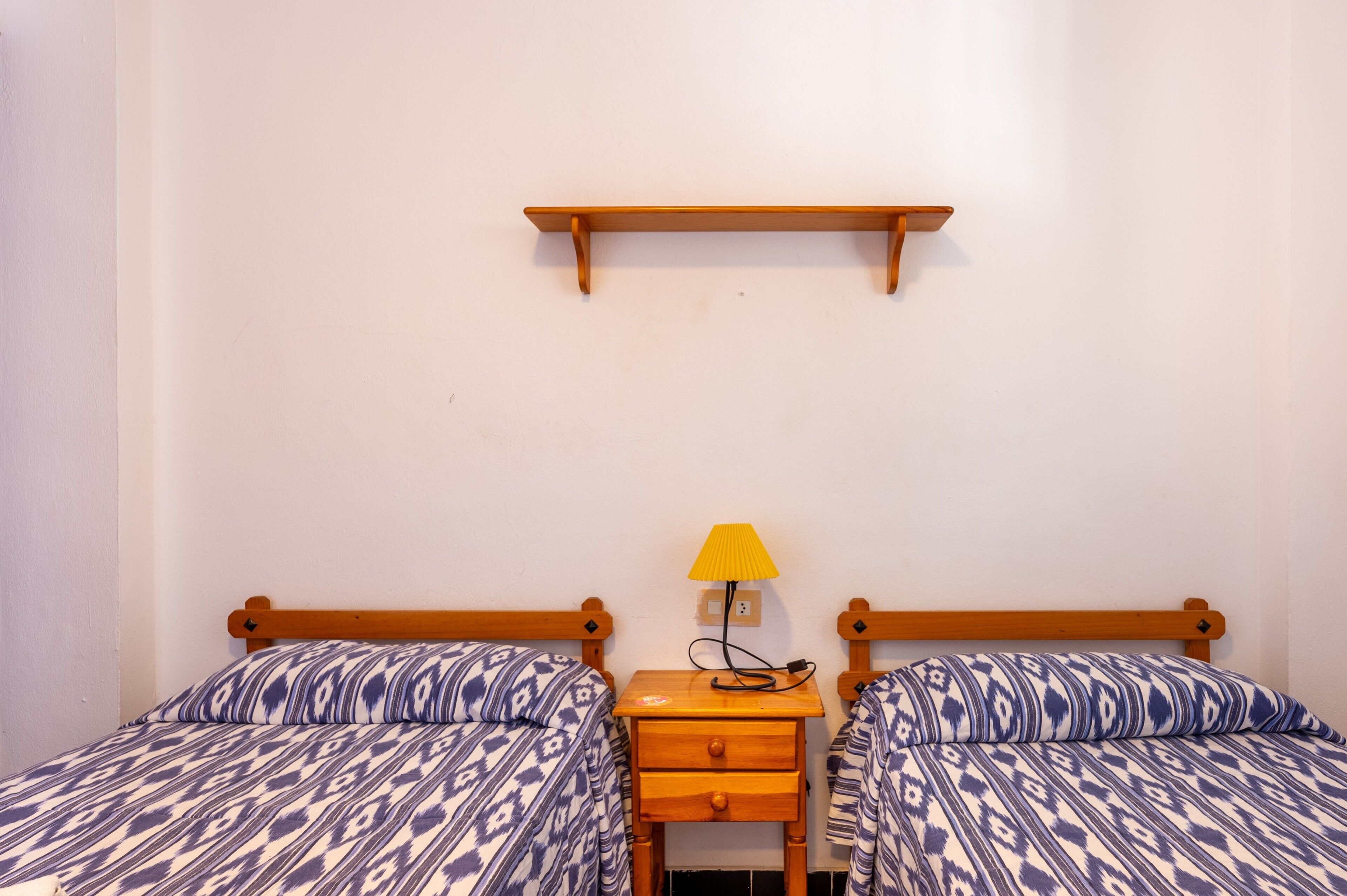 Una habitación con dos camas, una mesita de noche con lámpara y un estante de madera en la pared.