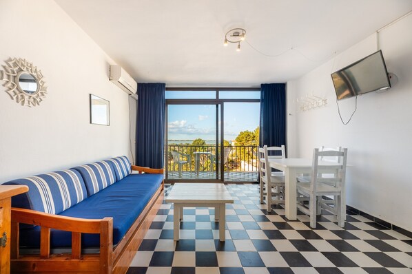 A room with a checkered floor, a sofa with striped cushions, a table, a TV mounted on the wall, and a balcony with chairs and a table. - Apartment 'Apartamento Bilo 303' with Lake View, Wi-Fi and Air Conditioning (Es Pujols)