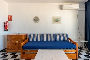 Ett rum med en blå soffa, ett träskåp, ett vitt bord, en brandsläckare, en väggmonterad luftkonditioneringsanläggning och en inramad tavla.