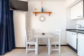 Une petite cuisine avec une table blanche, deux chaises, un téléviseur fixé au mur, une étagère avec un vase et un miroir sur le mur.