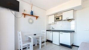 Una pequeña kitchenette con armarios blancos, un microondas y un lavamanos. Hay una mesa blanca con dos sillas y un televisor de pantalla plana montado en la pared.