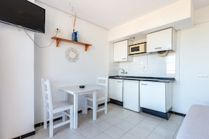 Una pequeña kitchenette con armarios blancos, un microondas y un lavamanos. Hay una mesa blanca con dos sillas y un televisor de pantalla plana montado en la pared.