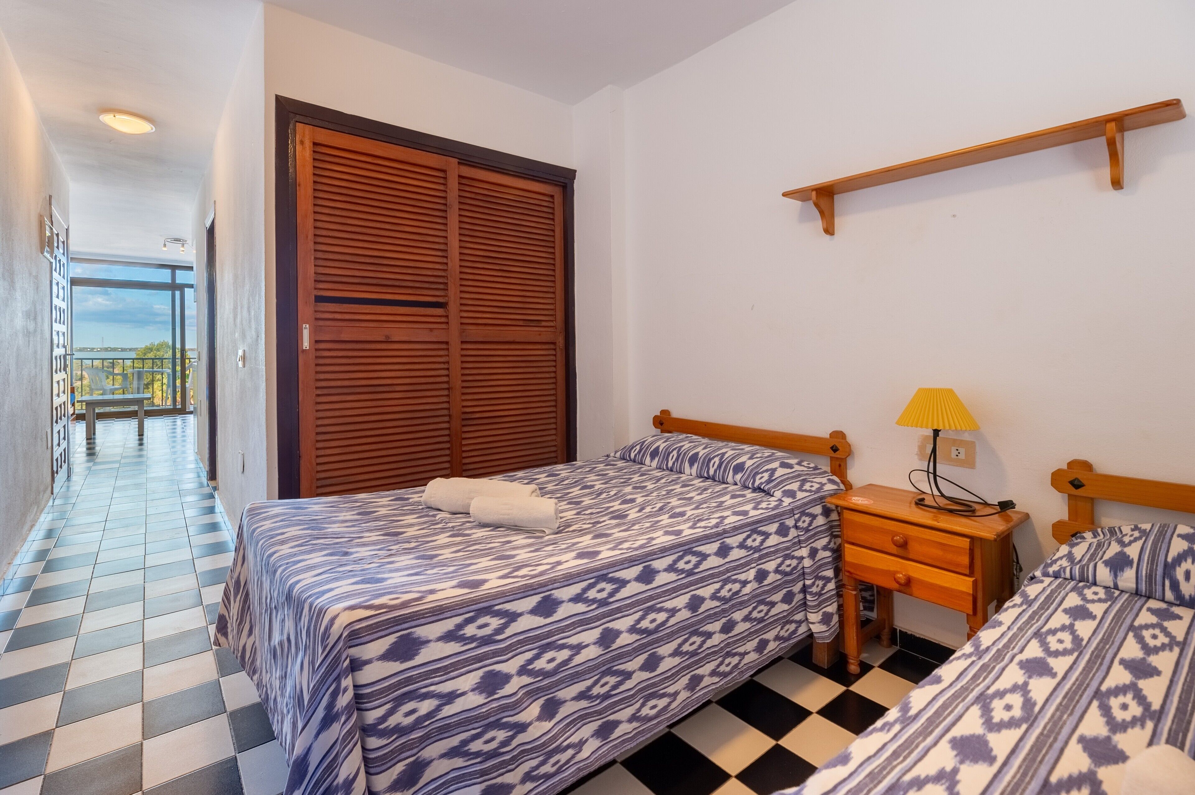 Una habitación de hotel con suelo a cuadros, una cama con ropa de cama estampada, una mesita de noche de madera con una lámpara y un armario grande de madera.