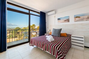 A hotel room with a large window, a bed with striped bedding, a nightstand, and two wall-mounted pictures. - Apartment 'Estudio Mirada 309' with Lake View, Wi-Fi and Air Conditioning (Es Pujols)