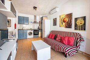 A compact living space with a kitchenette, a sofa, a white table, and artwork on the walls. - Apartment 'Trilo 24' with Wi-Fi and Air Conditioning (Es Pujols)