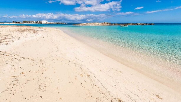 En strand med kristallklart turkosfärgat vatten, en sandstrand och byggnader i fjärran.