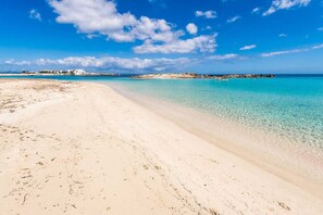 A beach with clear turquoise water, sandy shore, and buildings in the distance.