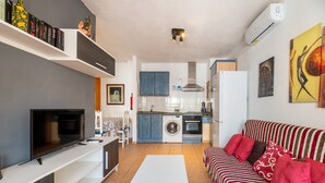 Ett modernt vardagsrum med en platt-TV, en soffa, ett soffbord och en köksdel med tvättmaskin.