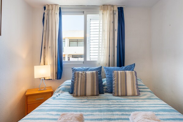 A bedroom with a bed, pillows, a nightstand, a lamp, and a window with curtains.